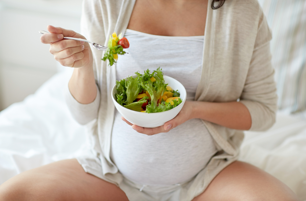 Pregnant woman eating bowl of salad in bed.